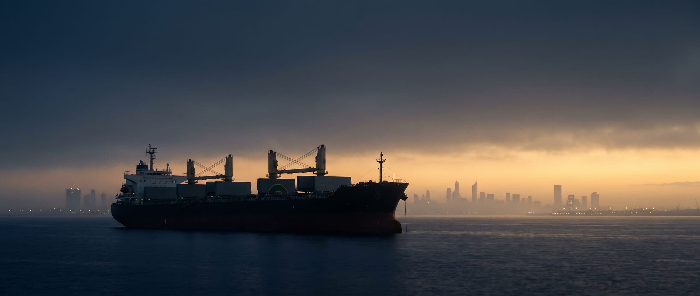 Bulk carrier at anchor at dawn before a city skyline